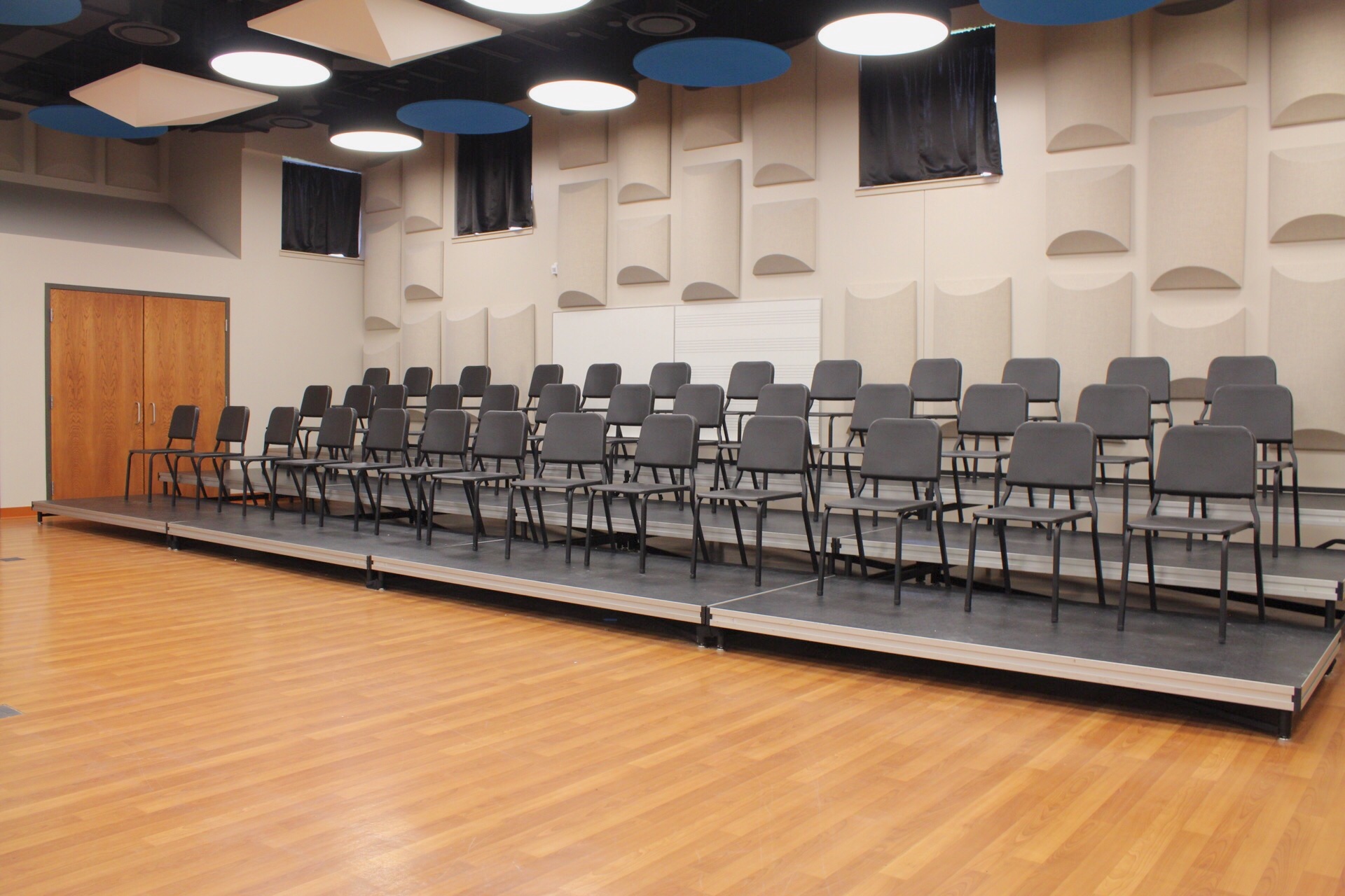 Jindra Fine Arts Choir Room has chairs on risers, black out curtains and acoustic panels on the walls. The chairs face a wall of mirrors.