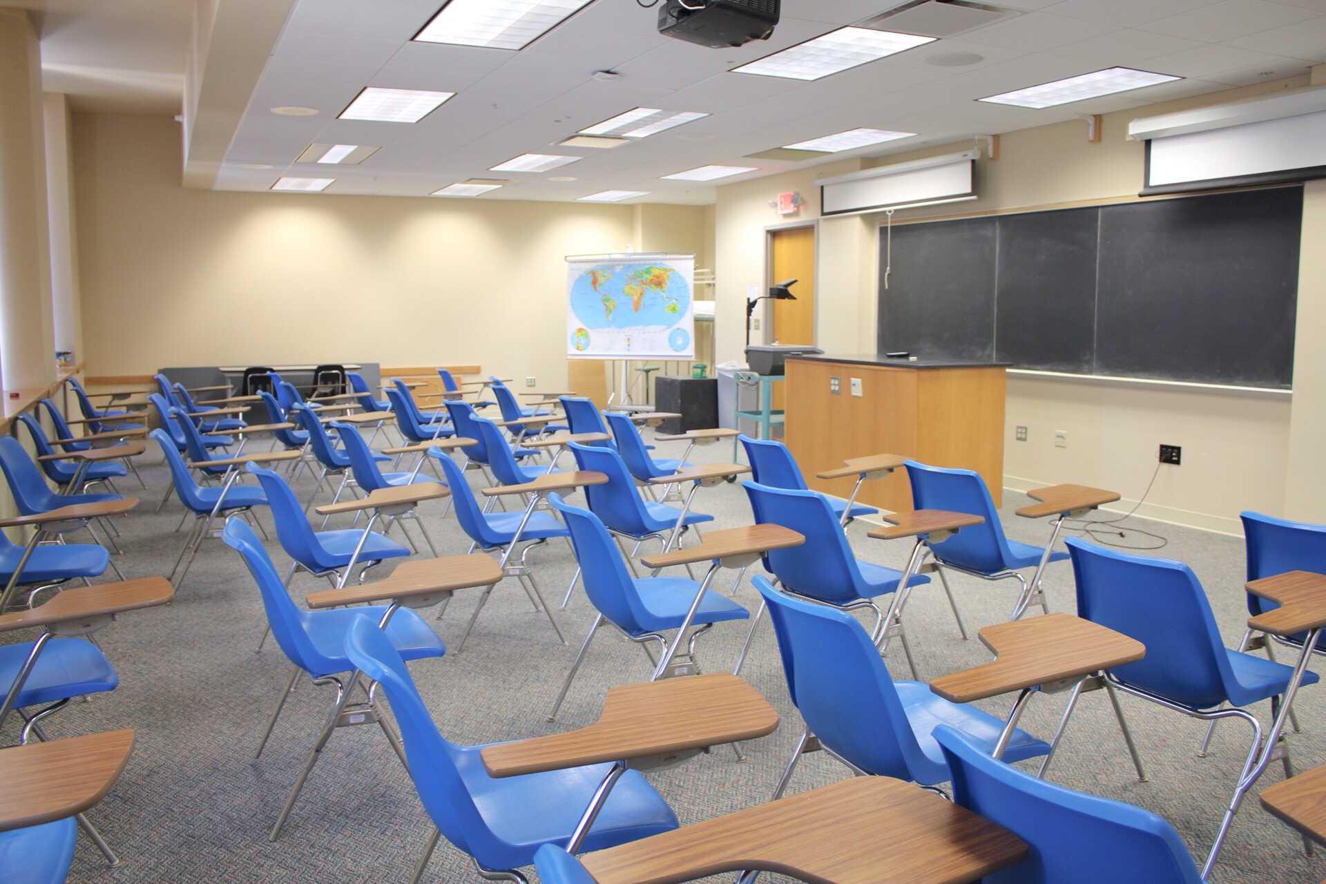 Hoyt Lecture 105 sideview of chairs with desk arms. a long chalkboard and 2 ceiling mounted projection screens.