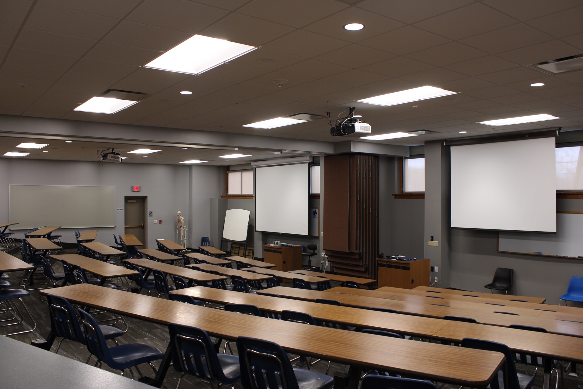 TJ Majors lecture 114 has 2 ceiling mounted projectors, 2 video screens, tables and chairs. The entire room can be split into 2 and the seated space is terraced.