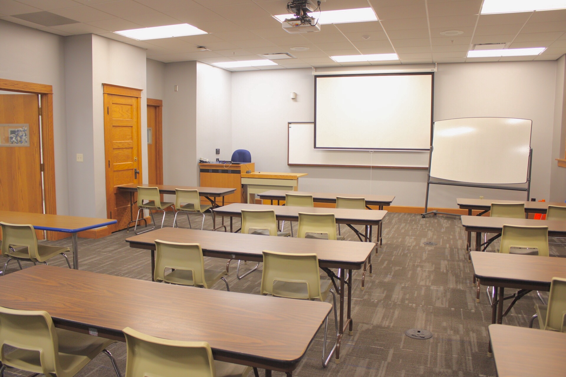 TJ Majors 301 has tables, chairs, a wall mounted white board, a movable white board, a ceiling mounted projector and a ceiling mounted projection screen.